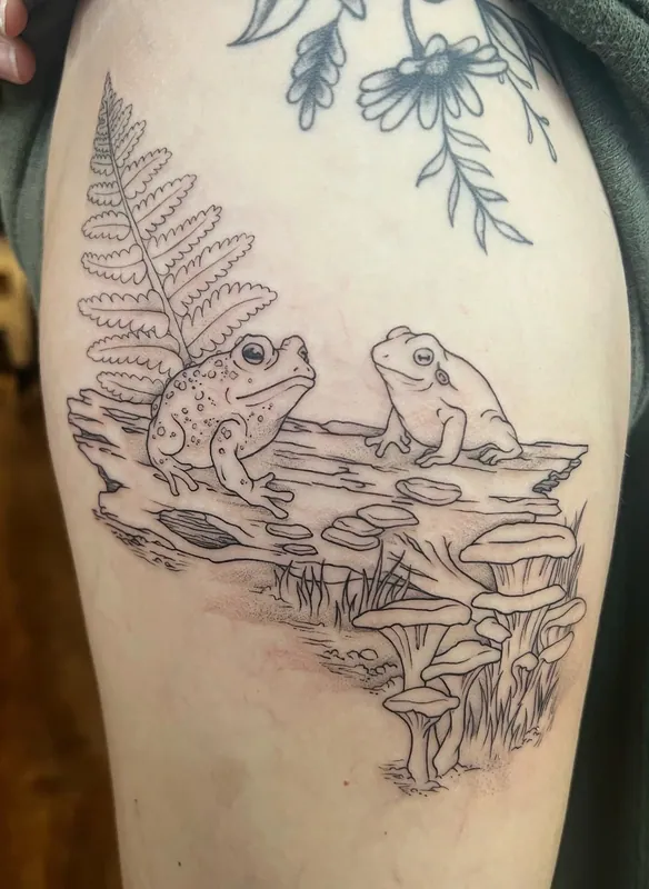 Fine-line tattoo of two frogs sitting on a log with a fern and mushrooms on an upper arm