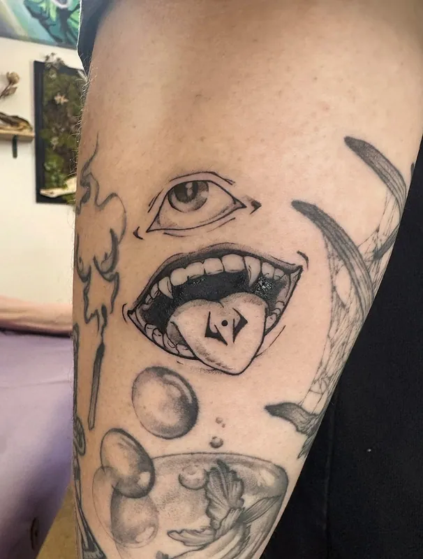 Black-and-grey surreal tattoo of an eye above an open mouth with a marked square on its tongue, bubbles and a fishbowl below, on an arm