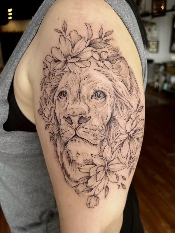 Fine-line black-and-grey lion portrait tattoo crowned and framed with magnolia blooms on an upper arm