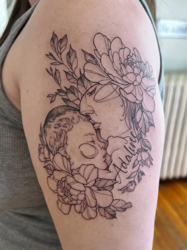 Fine-line black-and-grey tattoo of a mother kissing a sleeping baby's forehead, ringed by peonies and a script name, on an upper arm