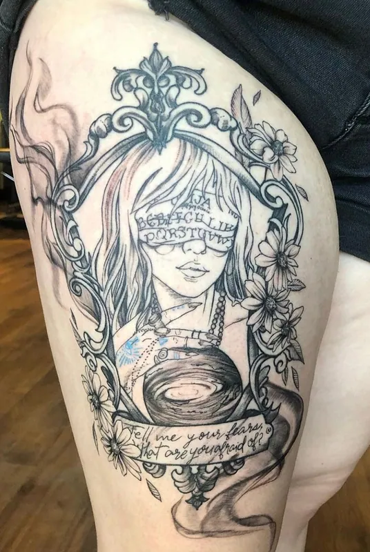 Black-and-grey tattoo of a woman blindfolded with lettered fabric, holding a galaxy, in an ornate frame with script below, on a thigh