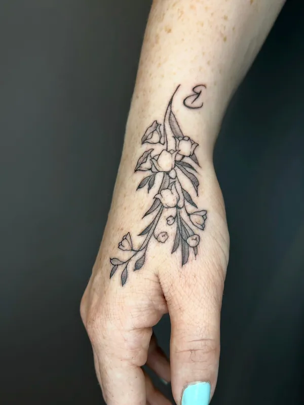 Fine-line black-and-grey tattoo of a tulip stem with leaves and an initial running from wrist onto the back of a hand
