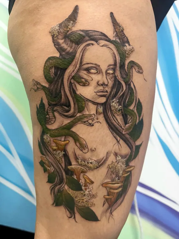 Color tattoo of a horned Medusa with green snakes in her hair and chanterelle mushrooms and lichen below on a thigh