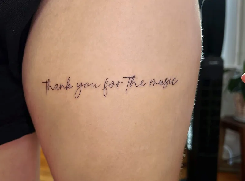 Fine-line cursive script tattoo reading 'thank you for the music' across a thigh