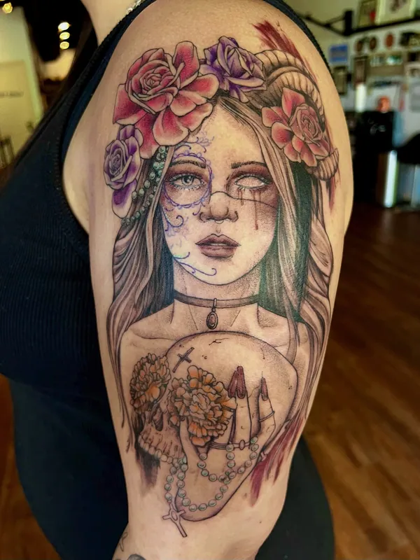 Color tattoo of a woman in sugar-skull face paint and a rose crown holding a marigold-filled skull and rosary on an upper arm