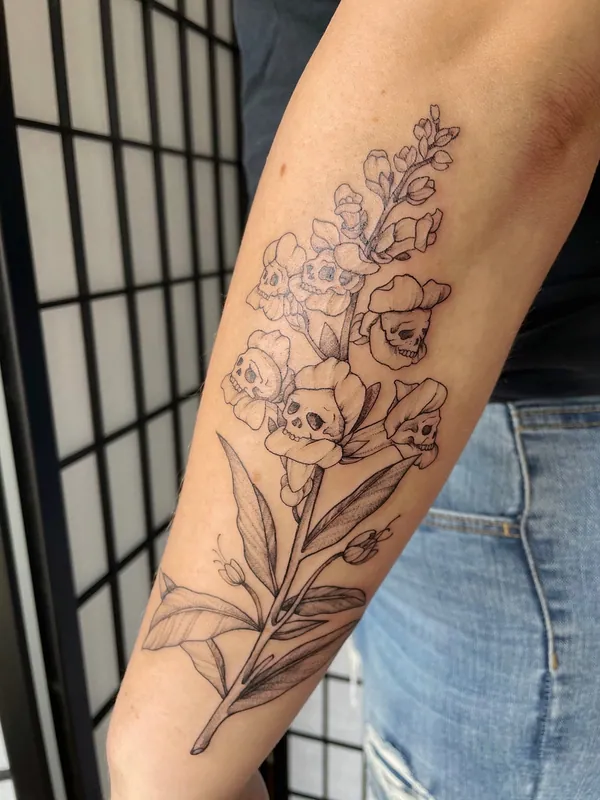 Fine-line black-and-grey snapdragon tattoo whose dried blooms form small skulls, on a forearm