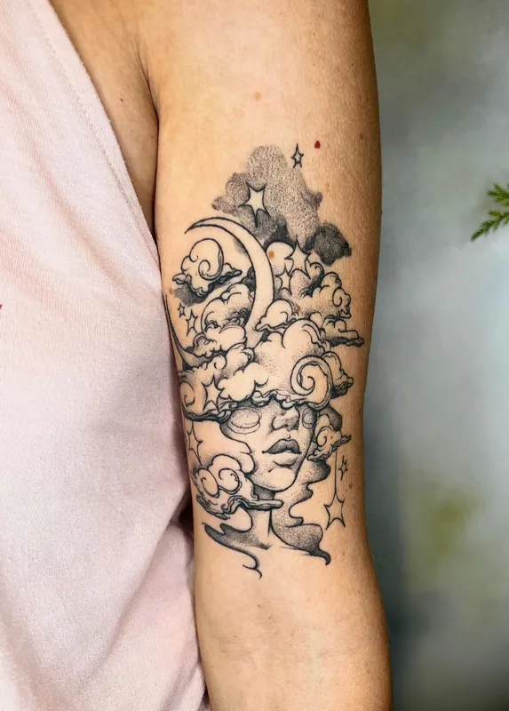 Black-and-grey dotwork tattoo of a face emerging from clouds beneath a crescent moon and stars on an upper arm