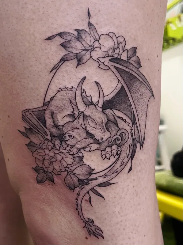Black-and-grey dotwork tattoo of a sleeping dragon curled inside a crescent moon with flowers on a thigh