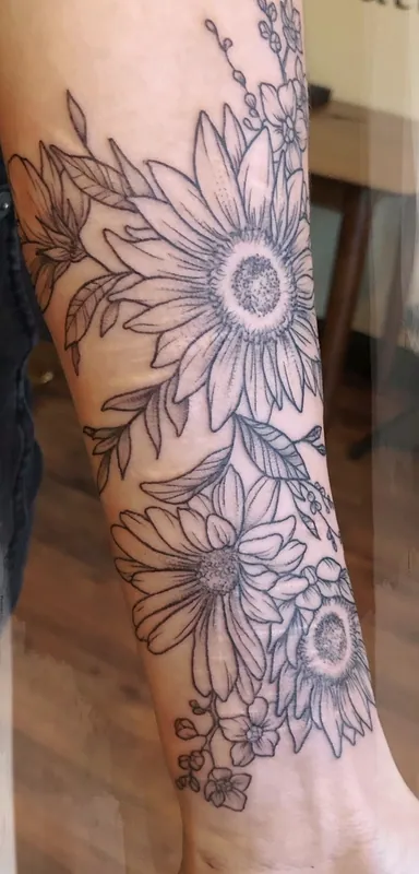 Black-and-grey fine-line tattoo of sunflowers and daisies with sprigs of wildflowers covering a forearm