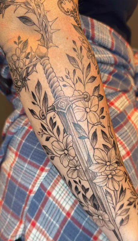 Black-and-grey fine-line sword tattoo wrapped in wildflowers and leaves along a forearm