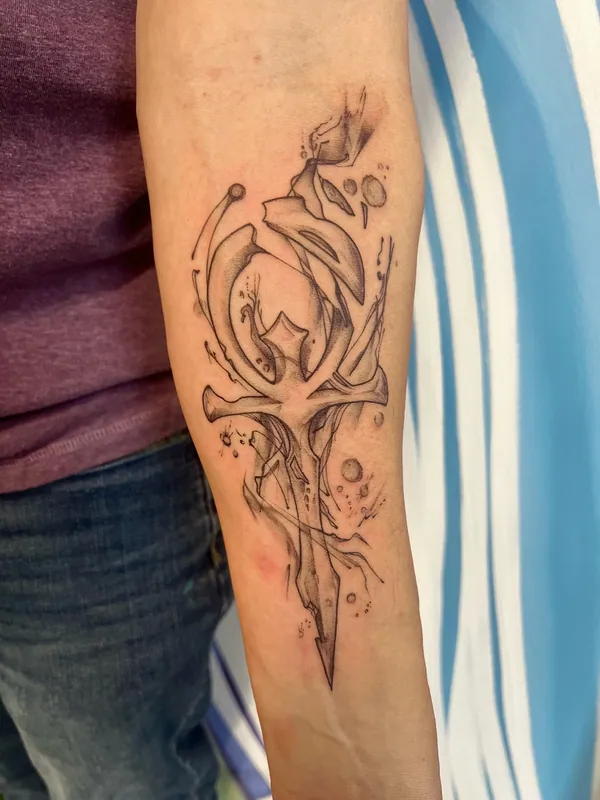 Black-and-grey tattoo of an ankh drawn as a blade with abstract smoke and ink spatter on a forearm