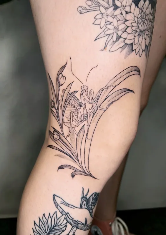 Fine-line black-and-grey praying mantis tattoo perched among grasses and wildflowers on a thigh