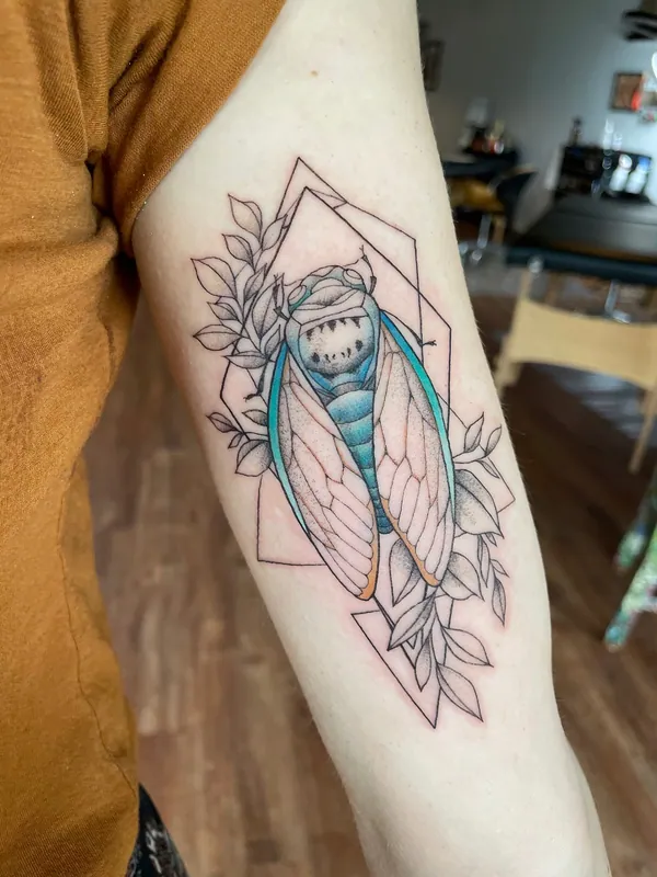 Fine-line cicada tattoo in teal color inside a geometric frame with leaves on an inner upper arm