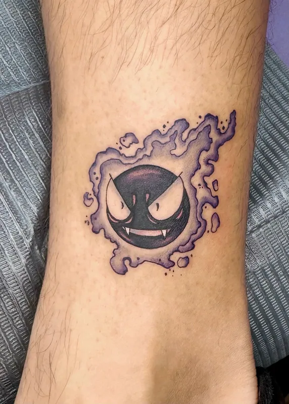 Color tattoo of Gastly from Pokemon wreathed in purple gas on a lower leg near the ankle