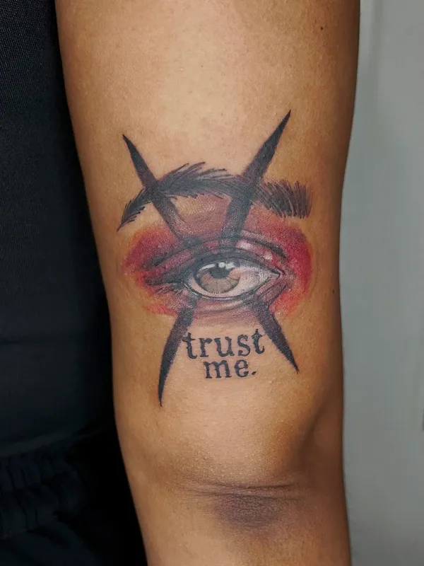 Color tattoo of an eye with red watercolor shading, crossed feather lines and the script 'trust me.' on an upper arm