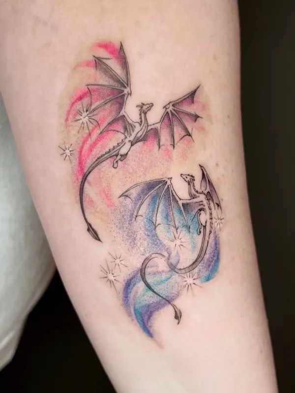 Fine-line tattoo of two flying dragons on red and blue watercolor washes with small stars on a forearm