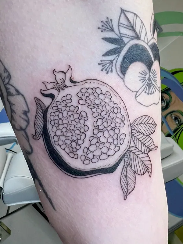 Linework tattoo of a halved pomegranate with seeds and hatched leaves on an upper arm