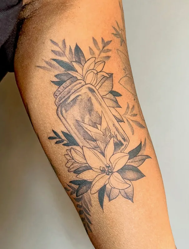 Black-and-grey fine-line tattoo of a mason jar holding a crystal, framed by lilies and leaves on an inner upper arm