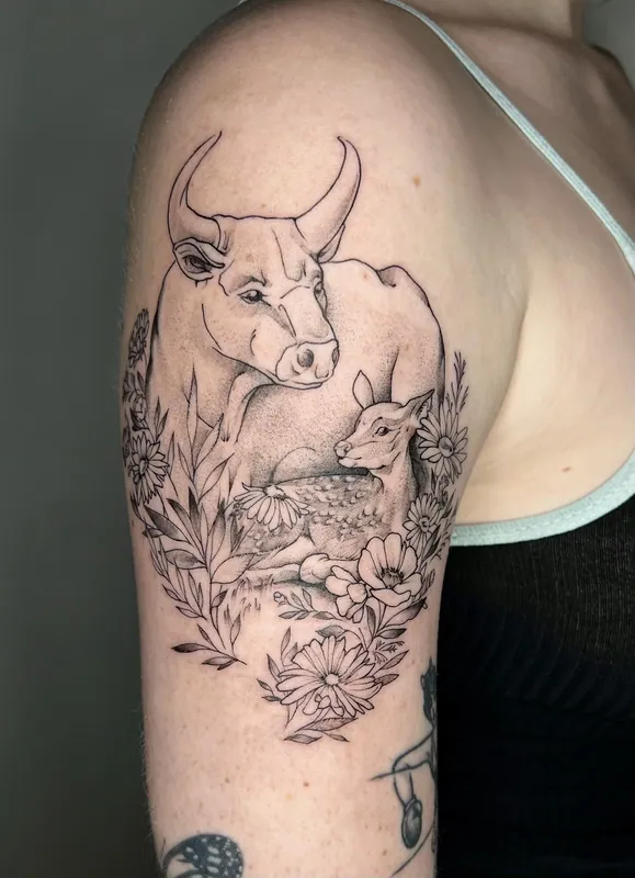 Fine-line black-and-grey tattoo of a horned cow standing over a resting fawn among daisies and poppies on an upper arm