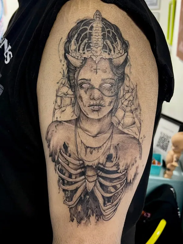 Black-and-grey tattoo of a horned figure with blank eyes, an inverted cross and an exposed ribcage on an upper arm