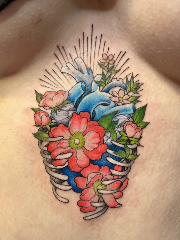 Color tattoo of a blue anatomical heart inside a ribcage with red and pink blooms and radiating lines on a sternum