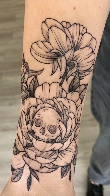 Black-and-grey fine-line tattoo of peonies with a small skull nested in the lower bloom on a forearm near the wrist