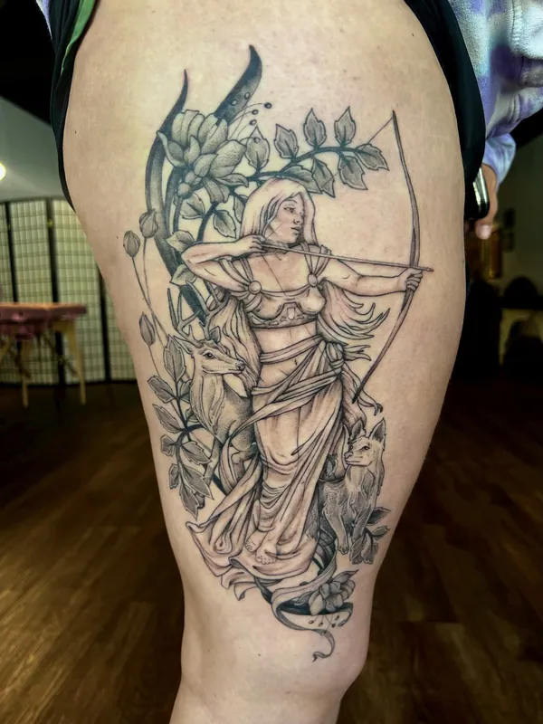 Black-and-grey tattoo of Artemis drawing a bow with a deer and hound at her side among leaves on a thigh