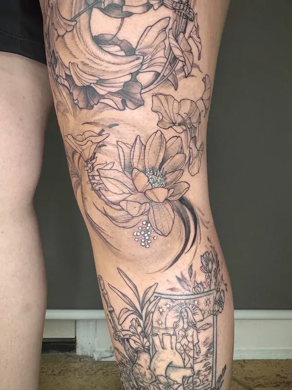 Black-and-grey fine-line lotus and botanical leg tattoo wrapping a knee, with a haloed figure below