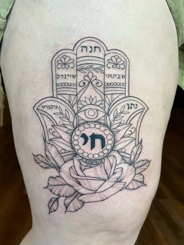 Black-and-grey hamsa tattoo with Hebrew lettering, a chai symbol, an eye and a rose at its base on a thigh