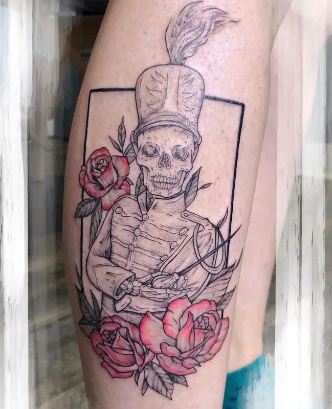 Tattoo of a skeleton in a plumed drum-major uniform with red color roses inside a thin frame on a calf
