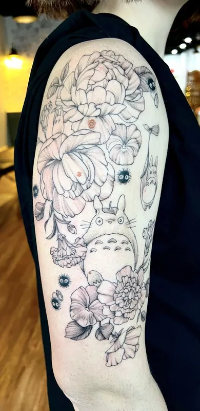 Fine-line black-and-grey Studio Ghibli tattoo of Totoro and soot sprites scattered among peonies on an upper arm