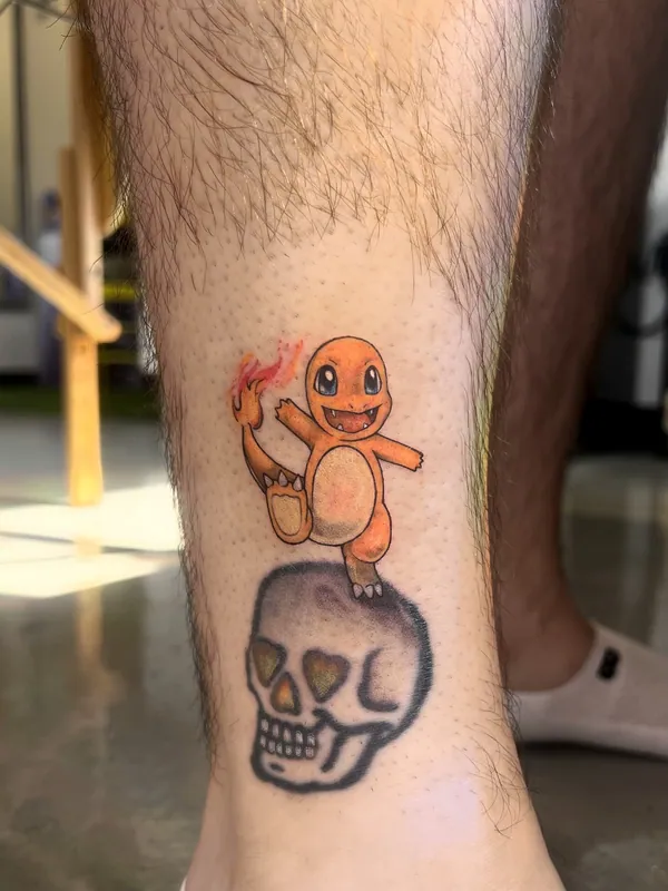 Color tattoo of Charmander from Pokemon standing on an older skull tattoo above an ankle