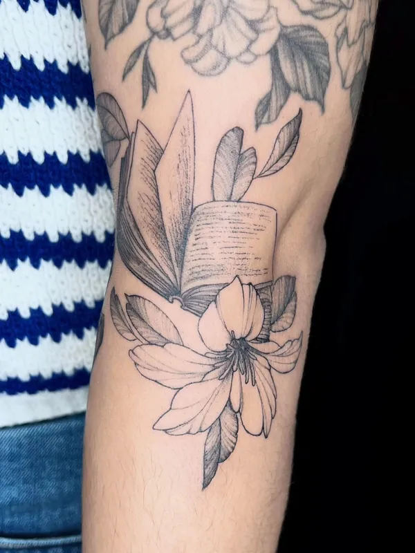 Black-and-grey fine-line tattoo of a lily blooming beneath an open book on an upper arm