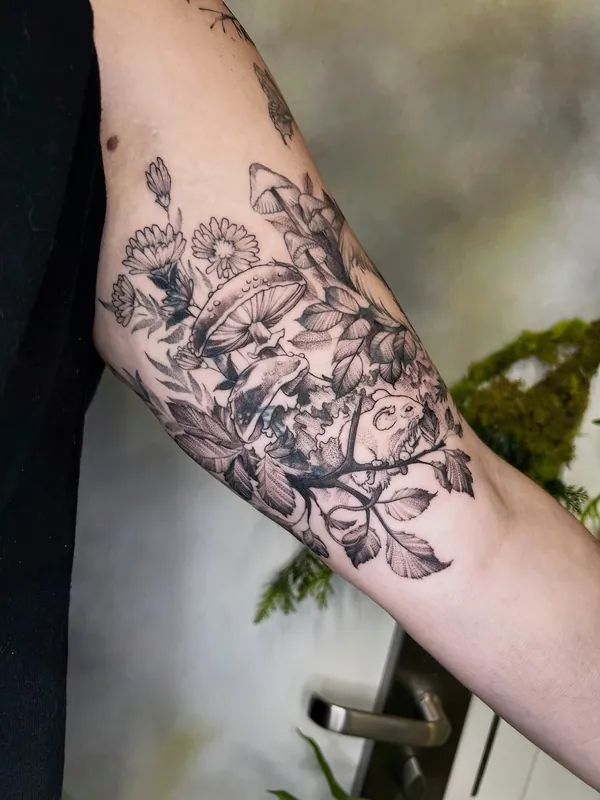 Black-and-grey fine-line tattoo of mushrooms, daisies and leaves with a small mouse on a branch on an inner upper arm