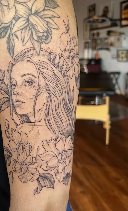 Fine-line black-and-grey tattoo of a woman's face framed by daffodils and blossoms on an upper arm