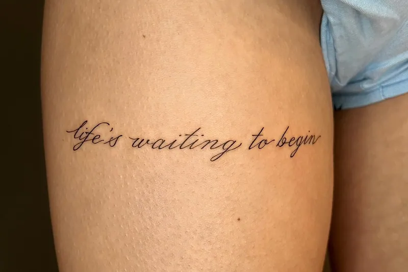Fine-line cursive script tattoo reading 'life's waiting to begin' across a thigh