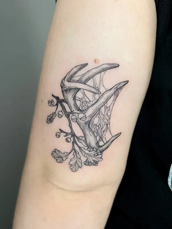 Fine-line black-and-grey tattoo of shed deer antlers with a spider web, oak leaves and acorns on the back of an upper arm