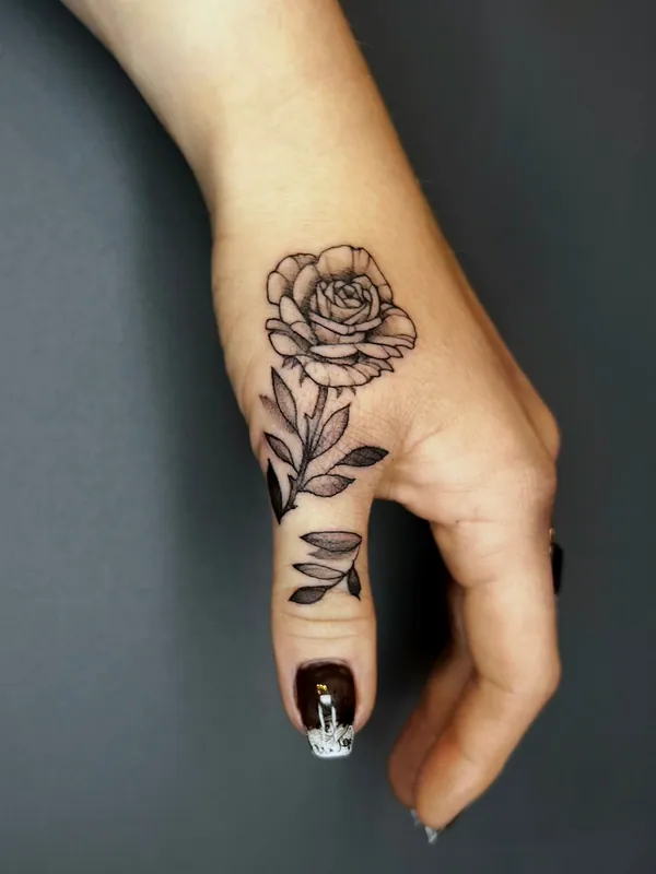Fine-line black-and-grey rose tattoo on the side of a hand with its stem and leaves running down the thumb
