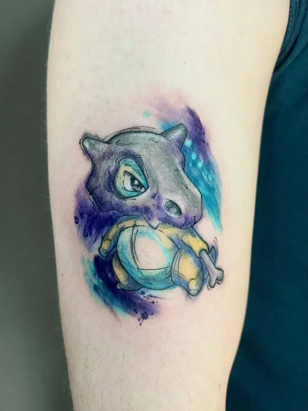 Color watercolor-style Cubone from Pokemon tattoo on a purple and teal wash on an upper arm