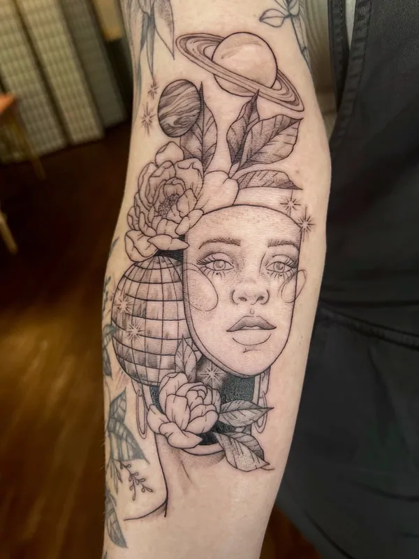 Black-and-grey tattoo of a woman's face beside a disco ball, with Saturn, a planet and peonies above, on a forearm