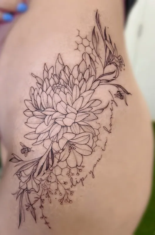 Fine-line chrysanthemum and lily tattoo with honeycomb, a bee and a line of script on a hip